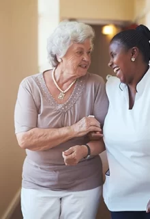 smiling older adult and her smiling caregiver walk down a hall together