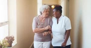 smiling older adult and her smiling caregiver walk down a hall together