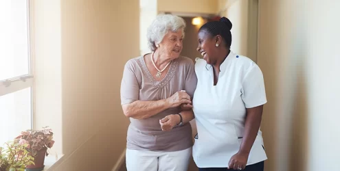 smiling older adult and her smiling caregiver walk down a hall together