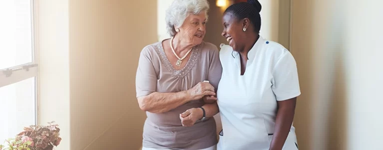 smiling older adult and her smiling caregiver walk down a hall together