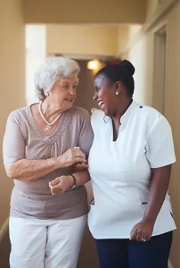 smiling older adult and her smiling caregiver walk down a hall together