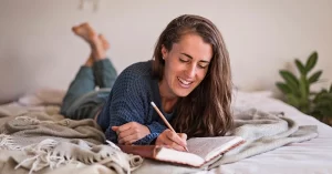 smiling woman lying on bed and writing in journal
