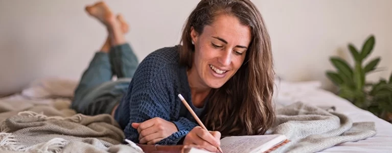 smiling woman lying on bed and writing in journal