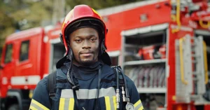 young black male firefighter stands in front of a firetruck