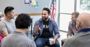 men sitting in a circle of chairs talking with an American flag behind them