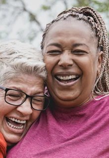 three older women stand outdoors in nature hugging and laughing