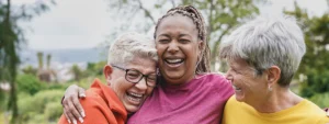 three older women stand outdoors in nature hugging and laughing