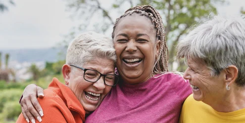 three older women stand outdoors in nature hugging and laughing