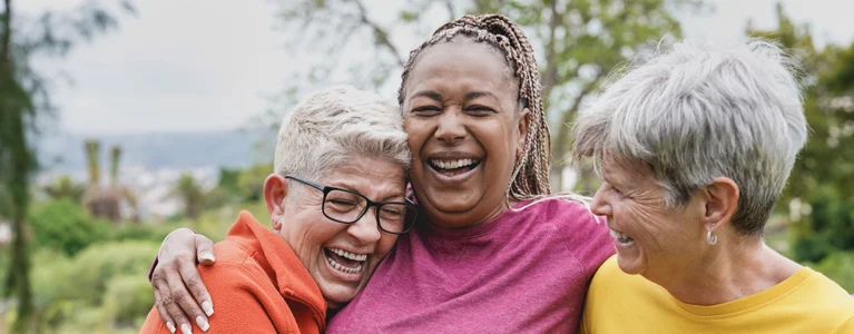 three older women stand outdoors in nature hugging and laughing