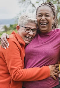 two older women stand outdoors in nature hugging and laughing