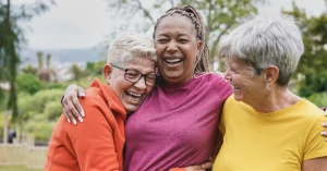 three older women stand outdoors in nature hugging and laughing