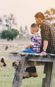 father and young son sit in a farm field looking at their sheep and goats