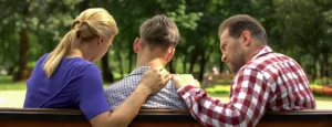 a teen and his parents sit on a park bench