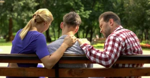 a teen and his parents sit on a park bench
