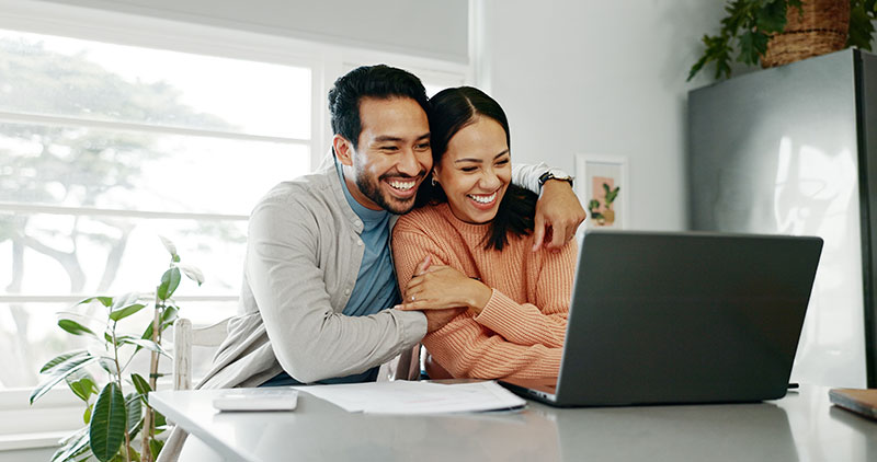 a man and woman sit together and hug in front of a laptop on a counter